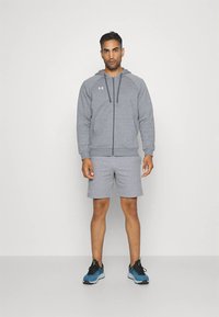 Vyras dėvi pilką užtraukiamą džemperį su gobtuvu ir atitinkančius šortus, mėlynais bėgimo bateliais, stovintis priešais baltą foną.