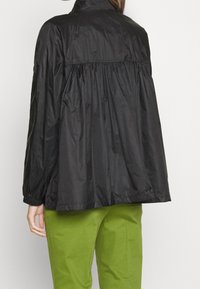 Czarna, lekka kurtka z zebranym tyłem, wysokim kołnierzem i elastycznymi mankietami; połączona z zielonymi spodniami. Teksturowany, wodoodporny materiał.