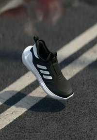 Sneaker atletico bianco e nero con tre strisce in volo sopra una strada asfaltata con segnaletica bianca.