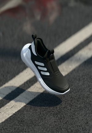 Sapatilha atlética em preto e branco com três riscas no ar acima de uma estrada asfaltada com marcas brancas de faixa.