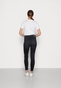 Vaqueros ajustados de cintura alta negros con detalles de costura sutil, combinados con una camiseta blanca y zapatillas blancas, vistos desde la parte posterior.