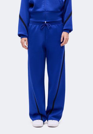 Personne portant un pantalon de sport bleu avec des bandes noires sur les côtés et des baskets blanches, debout devant un fond neutre.