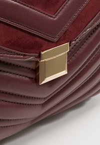 Sac à main en cuir bordeaux avec un design matelassé, des accents de quincaillerie dorée et une fermeture à boucle carrée. Texture douce en daim sur le dessus.