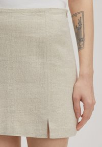 Beige textuur mini rok met een zijsluiting en een knoopsluiting. De stof heeft een subtiel, geweven patroon, wat een gestructureerde uitstraling creëert.