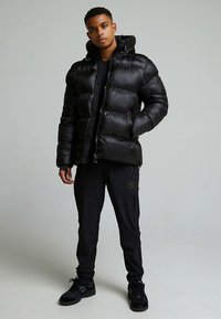 Μαύρο μπουφάν puffer με λαμπερό, καπιτονέ φινίρισμα, κουκούλα και εμπρόσθιο φερμουάρ. Συνδυάζεται με μαύρο παντελόνι και αθλητικά παπούτσια.