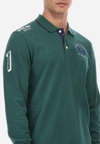 Zeleni polo majica z dolgimi rokavi iz bombaža, ima mornarsko modro ovratnik, izšito logo in podrobnosti številke na levem rokavu. Mehak otip.