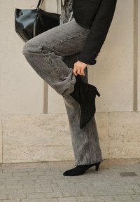 Svarta mocka ankelstövlar med spetsiga tår och en liten klack, kombinerade med blekta grå vida jeans. En enkel, texturerad svart handväska i bakgrunden.