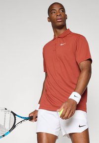 Polo košile v rezavé barvě s límečkem a logem Nike, spárované s bílými šortkami. V jedné ruce tenisová raketa a v druhé zelený tenisový míček.
