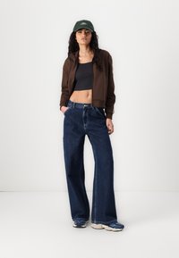 Brun zip-up jacka, svart crop top, vida mörkblå jeans och grå sneakers. Grön keps med vit text på framsidan. Lockigt hår synligt.