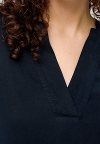 Granatowa bluzka z dekoltem w kształcie litery V i teksturowanym materiałem. Zbliżenie uwydatnia szczegóły szwów wokół kołnierza i ramion.