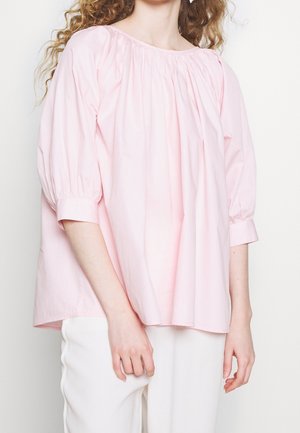 Kobieta w luźnej, jasnoróżowej bluzie z rękawami 3/4 o fasonie bufiastego, stojąca na tle jednolitego, jasnego tła i w białych spodniach.