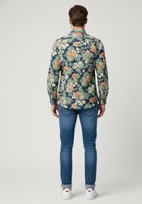 Camisa de botones con estampado floral que presenta flores verdes y naranjas sobre un fondo azul marino, combinada con jeans azul claro y zapatillas blancas.