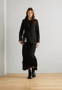 Blazer noir cintré sur une robe maxi noire, associé à des bottes noires à lacets. Le blazer présente deux poches avant et une fermeture à un bouton.