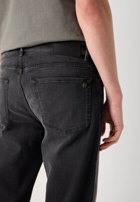 Gros plan d'une personne portant un jean en denim noir avec des poches arrière et une chemise grise, montrant le côté droit de la taille à la cuisse.