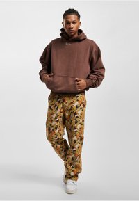 Sudadera grande marrón con bolsillo frontal y logo; pantalones cargo de camuflaje en tonos tierra; zapatillas blancas. Atuendo urbano informal.