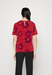Rød blomsterbluse med store lilla blomster og grønne blade, korte ærmer, rund hals med en lille nøglehul detalje bagpå.