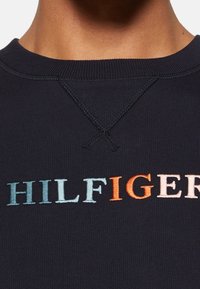Svart sweatshirt med ribbad halsringning, prydd med flersidiga broderade bokstäver som stavar "HILFIGER" i aqua, orange och persika.