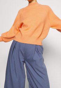 Suéter de punto naranja claro con cuello alto, hombros caídos y dobladillo acanalado, combinado con pantalones de traje azules de talle alto y con pliegues.