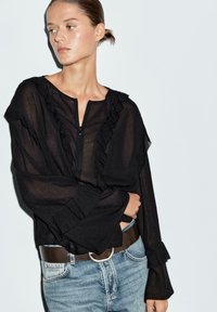 Svart genomskinlig blus med volangdetaljer och dragkedja i halsen, ihop med ljusblå jeans och ett brunt bälte med en cirkulär spänne.