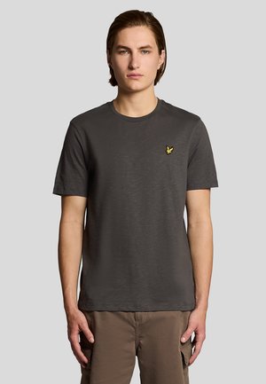 Jonge man met halflang haar draagt een donkergrijs T-shirt met korte mouwen met een klein geel adelaarlogo op de borst en een bruine broek.