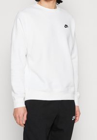 Bílá mikina Nike s kulatým výstřihem, žebrovanými manžetami a lemem. Na hrudi je malá černá značka. Kombinováno s černými kalhotami.