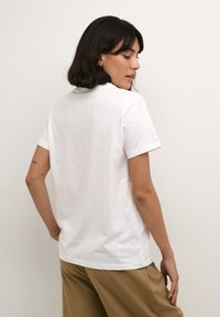 T-shirt en coton blanc à manches courtes et col rond, vue de l'arrière. Texture lisse, design simple, associé à un pantalon beige.