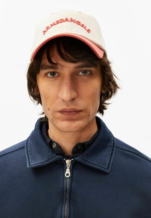 Giovane uomo con capelli castano scuro di media lunghezza che indossa un cappellino beige e rosa con la scritta "ARMEDANGELS" e una giacca blu navy con cerniera, che guarda direttamente davanti a sé.