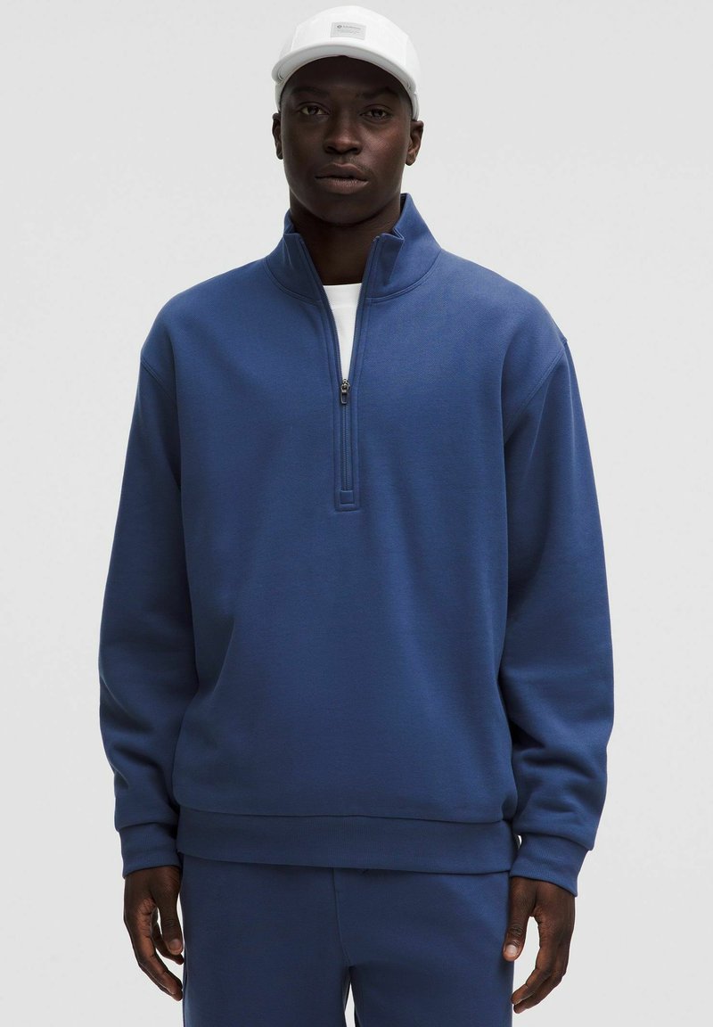 Felpa blu con zip a quarti, colletto alto, texture morbida e orlo a costine, abbinata a pantaloni blu coordinati.