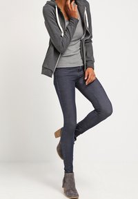 Sudadera gris con cremallera sobre una camiseta gris, combinada con jeans oscuros de corte slim y botines grises con tacón apilado. Textura suave en toda la prenda.