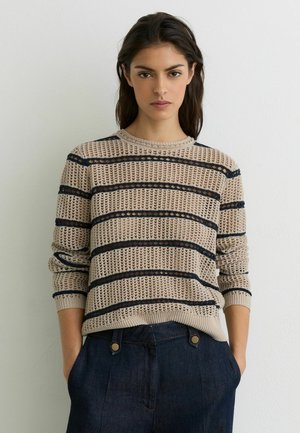 Kvinde iført en beige strikket trøje med vandrette sorte striber og mørkeblå bukser med høj talje, stående op ad en hvid væg.