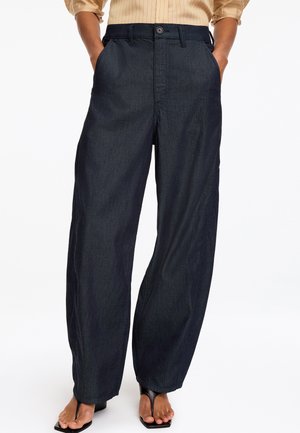 Person trägt dunkelblaue High-Waist-Jeans mit weitem Bein, die Hände in den Taschen, und schwarze offene Sandaletten mit Absatz vor weißem Hintergrund.