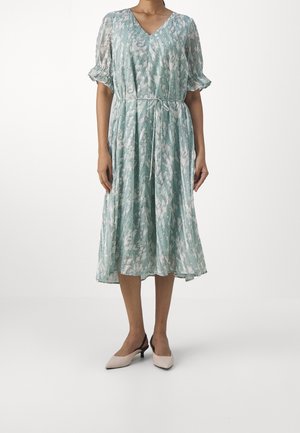Frau trägt ein hellblau-weiß gemustertes Midikleid mit kurzen Puffärmeln und einem Taillengürtel, kombiniert mit beigen Slingback-Pumps mit niedrigem Absatz.