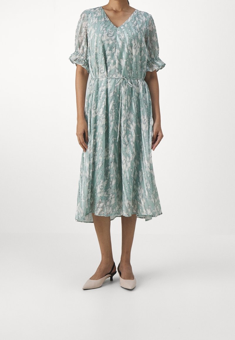 Femme portant une robe midi à motif bleu clair et blanc avec des manches courtes bouffantes et une ceinture à la taille, associée à des chaussures beige à talons bas et bride arrière.