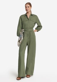 Olivgrön overall med långa ärmar, kragdesign, två bröstfickor, bälte i midjan och en mönstrad scarf knuten i bältet.