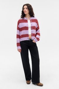 Donna che indossa un cardigan felpato a righe viola e ruggine, un top corto bianco, pantaloni neri larghi e stivali marroni, in piedi con una mano in tasca.