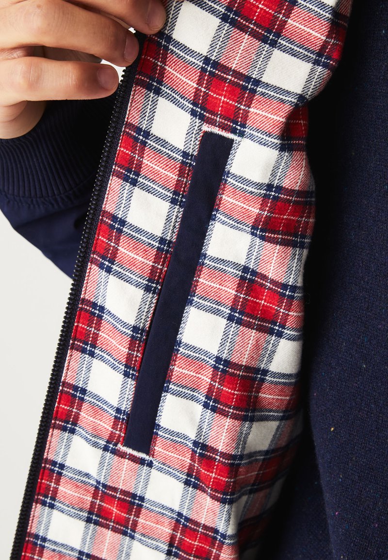 Intérieur d'une veste zippée marine avec une doublure en tartan blanc, rouge et bleu ; présente une texture lisse et une poche cachée avec un accent marine.