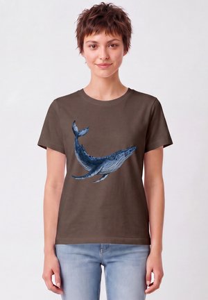 Jonge persoon met kort bruin haar, draagt een bruin T-shirt met een blauwe walvisillustratie en lichtblauwe jeans, staand tegen een effen achtergrond.
