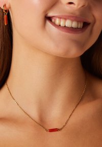Gouden ketting met een rechthoekige oranje emaille hanger, gecombineerd met bijpassende oranje ringoorbellen. Glad oppervlak, modern ontwerp.