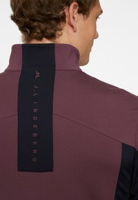 Lila träningsöverdel med hög krage, som har en svart vertikal panel på baksidan med präglad logotyp. Slät tygkvalitet.