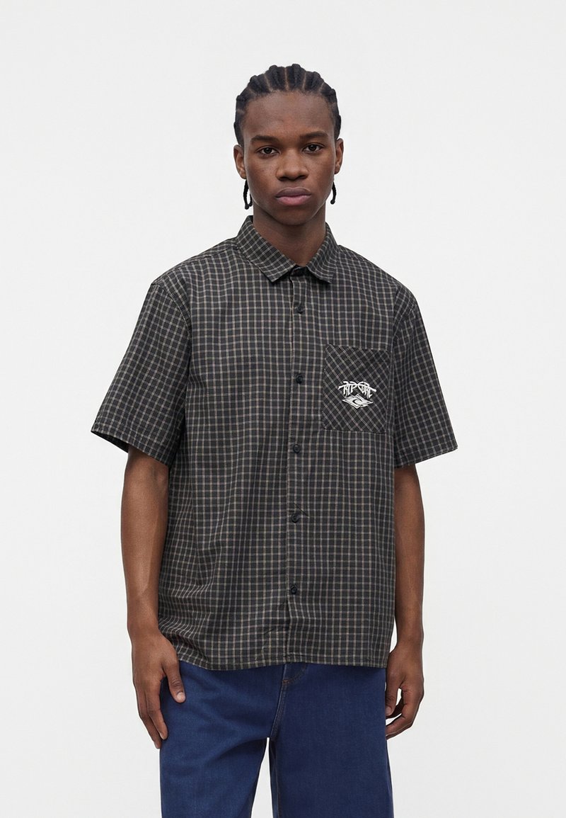Jeune homme aux cheveux tressés porte une chemise à manches courtes à carreaux noirs et gris avec une poche avant et un jean bleu, debout devant un fond uni.