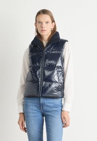 Gilet imbottito blu navy in materiale lucido e trapuntato, con un colletto morbido in pile. Presenta una zip frontale e due tasche laterali. Indossato sopra una camicia bianca.