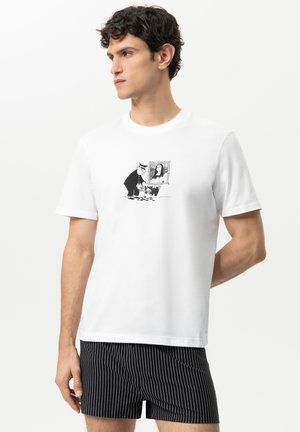 Mann trägt weißes T-Shirt mit einem schwarz-weißen Cartoon eines Mannes, der Pflanzen gießt, unter einem gerahmten Mona-Lisa-Gemälde, kombiniert mit schwarzen Nadelstreifen-Shorts.