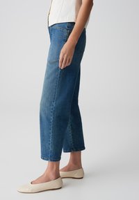 Högmidjade, raka blå denimjeans med en ljus tvätt, subtil blekning och uppvikta avslut, tillsammans med beige platta skor.