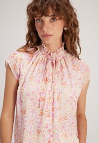 Bloemmotief blouse in lichtroze met oranje en paarse patronen. Rimpelnek, korte mouwen en knoopsluiting aan de voorkant. Zachte stoftextuur.