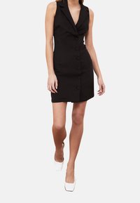 Robe noire sans manches avec un décolleté en V profond, un design structuré, des boutons à l'avant et une silhouette ajustée, associée à des talons blancs à bout carré.