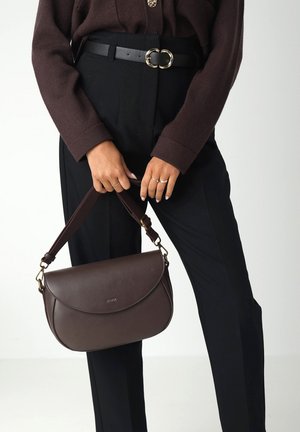 Borsa a tracolla in pelle marrone con design arrotondato, tracolla sottile e regolabile, e dettagli in oro. Abbinata a pantaloni neri a vita alta e un top marrone.
