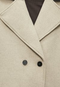 Διπλής όψης καφαντάρι σε μπεζ από υφασμάτινη υφή, με φαρδύ γιακά και δύο μαύρα κουμπιά μπροστά.