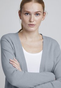 Gai pelēks adīts kardigans ar V-veida kaklu, pilnām piedurknēm un ribotām aprocēm, valkāts virs baltas T-krekla. Audums izskatās mīksts ar smalku tekstūru.