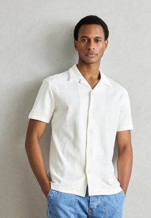 Weißes Kurzarmhemd aus strukturiertem Stoff, mit einem Notch-Kragen und sechs Knöpfen auf der Vorderseite. Getragen mit blauen Jeans-Shorts.