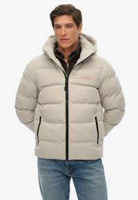 Chaqueta acolchada beige claro con capucha, cremallera negra y bolsillos laterales. Diseño acolchado con una textura suave. Lleva una camisa oscura debajo.